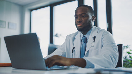 Serious African American Family Medical Doctor is Working on a Laptop Computer in a Health Clinic. Physician in White Lab Coat is Browsing Medical History Behind a Desk in Hospital Office. 