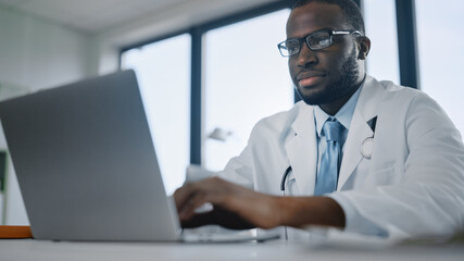 Happy African American Family Medical Doctor is Working on a Laptop Computer in a Health Clinic. Physician in White Lab Coat is Browsing Medical History Behind a Desk in Hospital Office. 