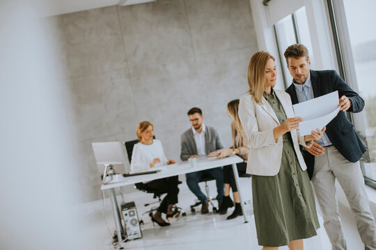 Man And Woman Discussing With Paper In Hands Indoors In The Office With Young People Works Behind Them