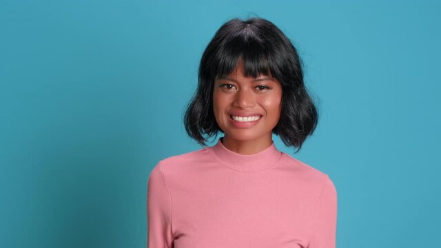 Silly cute dark skinned woman shrugs slightly and smiles looking at camera. Young glamorous female in pink blouse looks clueless standing on blue background. Girl has no idea about the situation.