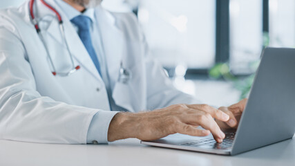 Close Up of a Family Medical Doctor Working on a Laptop Computer in a Health Clinic. Physician in White Lab Coat is Browsing Medical History Behind a Desk in Hospital Office. 