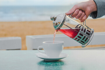 Man in blue jacket pouring tea in mug at cafe at beach on seaside yellow sand. Steel metal tea pot with red fruit tea and white tea cup. Concept of rest at sea, relaxation, vacation, peace.