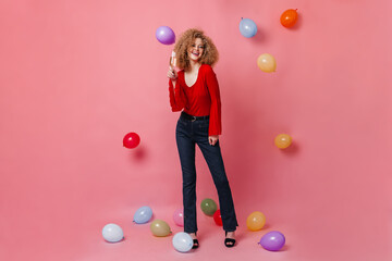 Fototapeta premium Cheerful girl with glass of champagne posing on pink background among balloons. Snapshot of curly woman in red top and denim pants