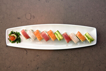 Assortment of Japanese sushi rolls nigiri, sashimi, maki, salmon, tuna and avocado served in long rectangular plate over stone background, top view