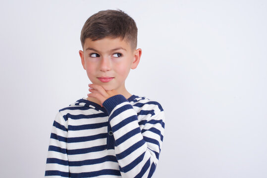 Dreamy little cute boy kid wearing red stripped t-shirt against white wall with pleasant expression, looks sideways, keeps hand under chin, thinks about something pleasant.