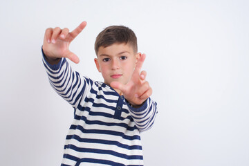 Portrait of smiling little cute boy kid wearing red stripped t-shirt against white wall looking at camera and gesturing finger frame. Creativity and photography concept.