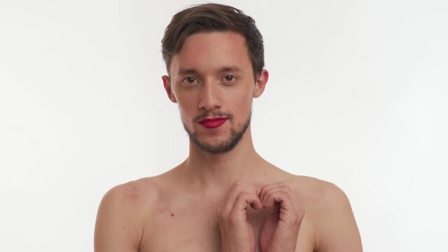 Beautiful young half naked Caucasian brunette bearded man with brown eyes wear makeup &ndash; red lipstick on lips smile, make gesture heart with hands, blow kiss, stare at camera on white background close 