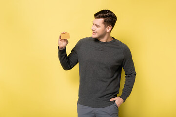 Smiling man with hand in pocket looking at credit card on yellow background