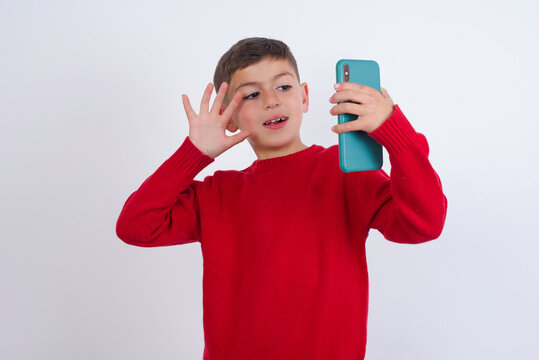 Portrait of happy friendly Little cute boy kid wearing red knitted sweater against white wall taking selfie and waving hand, communicating on video call, online chatting.