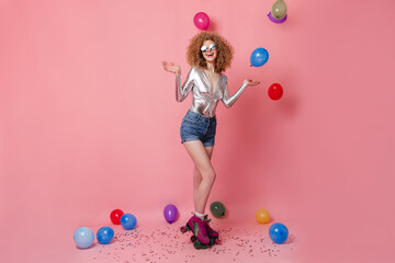 Adorable girl in sunglasses, silver blouse and shorts roller skating in pink studio among multicolored balloons