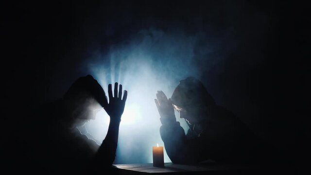 Two men in hoods sit by a burning candle, behind them shrouded in fog illuminated by a spotlight