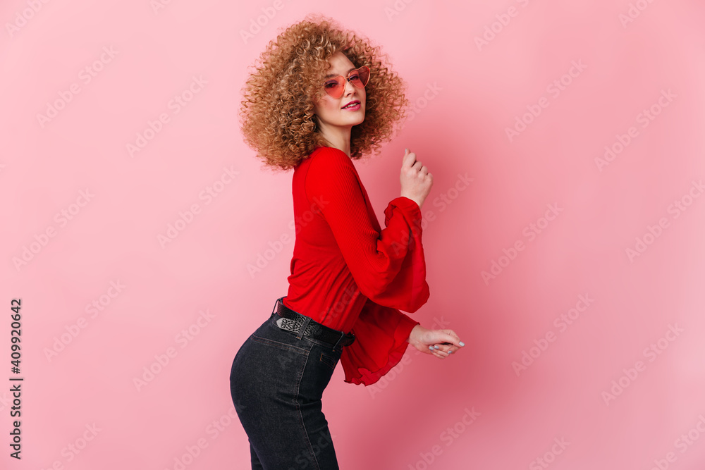 Wall mural Attractive blonde girl in sunglasses dressed in top with wide sleeves and dark pants dancing on pink background