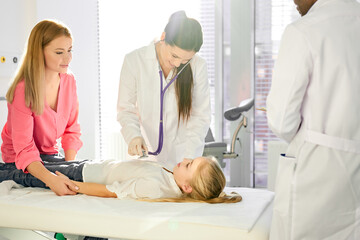 Obraz premium caucasian female doctor check the heartbeat of patient child with stethoscope while his african male colleague help her, in modern clinic