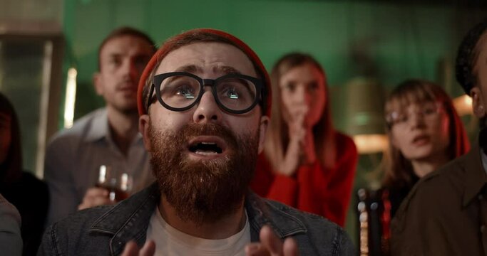 Close up view of bearded guy in glasses watching sports game with friends in pub and looking upset. Group of multiethnic people become sad while team they supported lose