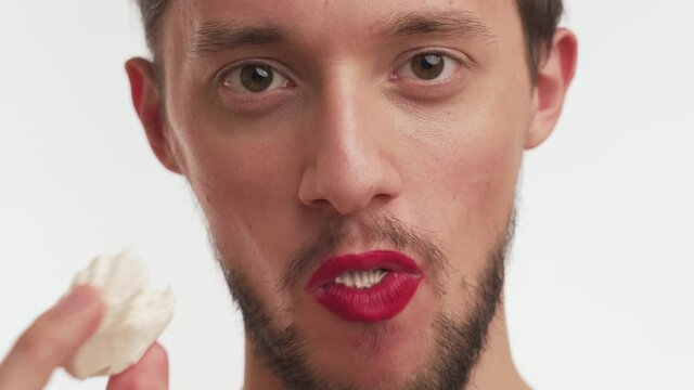 Close up portrait of Caucasian transvestite male eating. Sexy man with black beard, painted lips in red color lipstick stare at camera, open mouth wide, put delicious marshmallow, chews with pleasure.