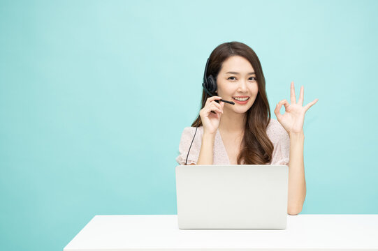 Young Asian Woman Friendly Operator Agent With Headsets And Showing Ok Sign Working In A Call Centre Department Isolated On Light Green Background