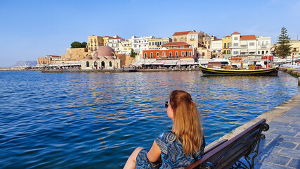 Fototapeta na wymiar A young woman sits on a bench at The Old Venetian Harbor of Chania, Crete