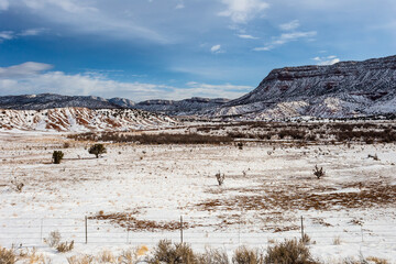 Large open snow covered field with vintage barbwire fence in front of a large mesa plateau with clouds in sky in rural New Mexico