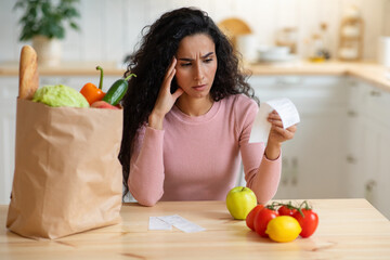 Grocery Expenses. Concerned Woman Sitting In Kitchen, Looking At Food Shopping Bill