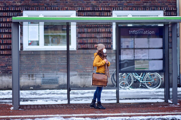 Midde-aged brunette woman standing at a bus stop, wearing a protective face mask due to corona virus, waiting for her bus to bring her to work