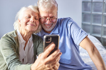 elderly couple taking photo on smartphone, while sitting in bedroom, sit smiling. senior people society lifestyle technology concept. man and woman share social media together in wellbeing home