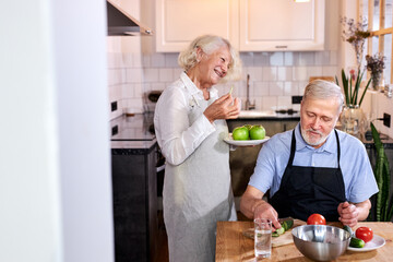 Fototapeta na wymiar elderly couple in the kitchen at home, senior man in apron sits carving fresh vegetables, gray-haired woman holding apples in hands and talking with him