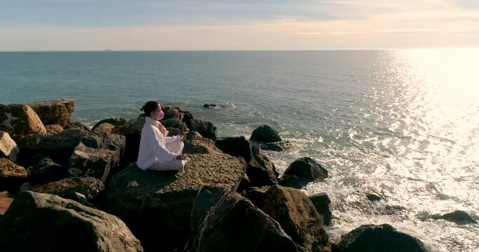 Mujer sentada postura de yoga meditaci&oacute;n con mascarilla de protecci&oacute;n junto al mar