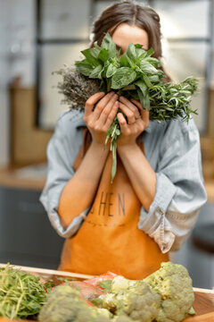 Portrait of cheerful woman in apron holding fresh spicy herbs basil, rosemary, thyme and cover her face. On the kitchen. Healthy cooking concept. High quality photo