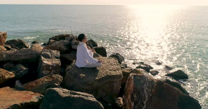 mujer vestida de blanco meditando ojos cerrados sobre rocas en mediterr&aacute;neo
