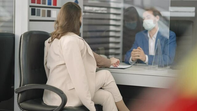 Back View Of Young Woman Signing Documents In Bank Or Organization On Covid-19 Pandemic With Employee Sitting Behind Glass In Face Mask. Business Lifestyle On Coronavirus Lockdown. Slow Motion.