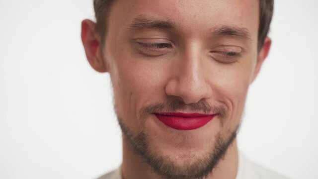 Young cute transvestite embarrassed brunette man with dark black beard, moustache, painted lips in red lipstick rolls eyes, looks around, stare at camera, smiles isolated on white background close up.
