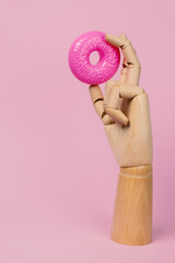 Wooden hand with pink donut on a pastel pink background. Fake plastic food and unhealthy diet concept.