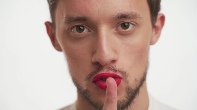 One young handsome shy Caucasian brunette man with brown eyes, beard, mustache puts finger to painted lips by red lipstick, show gesture shh silence or secret isolated on white background close up.