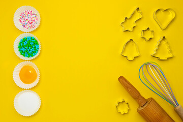 Ingredients, sugar decoration, kitchen tools for baking on the yellow background. Cookie cutters, a rolling pin, an egg whisk. Yolk, sugar, baking decoration in muffin forms. Copy space. Top view.