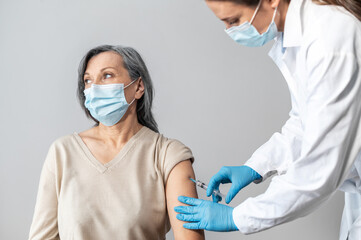 An elderly woman getting a vaccine for coronavirus, a doctor wearing mask and gloves with a syringe...