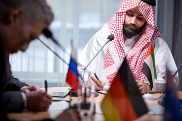 serious sheikh in traditional wear sits signing document, at business meeting in the office, concentrated