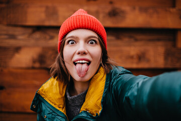 Close-up portrait of perky teenager girl taking selfie on wooden background. Model in warm jacket and hat shows tongue