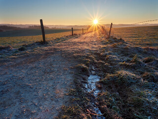 Frosty sunrise in the German city of Wipperfurth. A beautiful countryside landscape in the Bergisches Land region. A field road covered with frost in the rays of the rising sun.