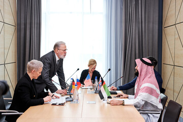 Fototapeta premium senior politician offers his plan of action and share opinion, elderly man in suit talking to multi-ethnic group of partners sitting at desk in boardroom, discussing