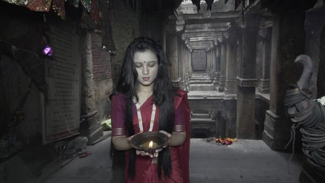 beautiful Indian girl in old stepwell wearing traditional Indian red saree, gold jewellery and bangles holding fire plate. Maa Durga shoot concept. Indian girl dancer in the posture of Indian dance