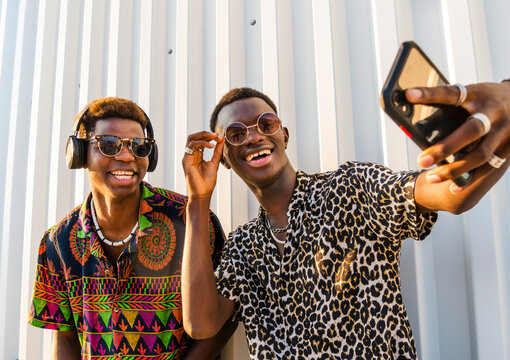 Optimistic African American male friends in fancy outfit and sunglasses taking selfie on smartphone and enjoying weekend on street