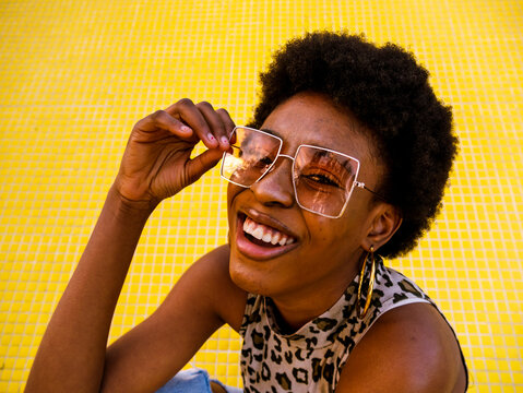 From above of optimistic young African American female in trendy ripped jeans and stylish sunglasses sitting on yellow floor and laughing happily looking at camera