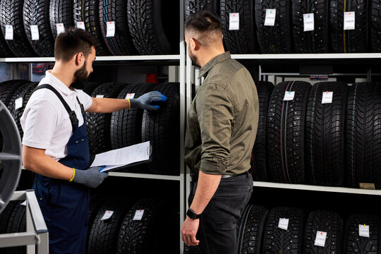 The Seller Offers New Rubber Wheels, Tires, Rims For The Car. Male Client Came To Make Purchase In Auto Service Shop. Auto Mechanic Showing Assortment Of Tires