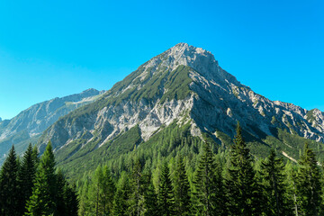 Fototapeta premium A clear view on Mittagskogel in Austrian Alps. The mountain stands independently. The high mountain is overgrown with a lush forest, with barren and sharp peak. Clear and sunny day. Alpine landscape