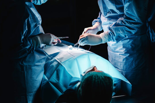 Crop anonymous surgeon and assistant with surgical tools and thread stitching wound of patient during operation in hospital
