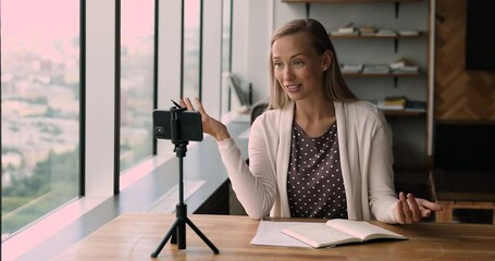 Skilled 30s female business coach influencer blogger filming educational workshop or lecture on mobile front camera in office, sitting at workplace table, caucasian woman making online presentation.