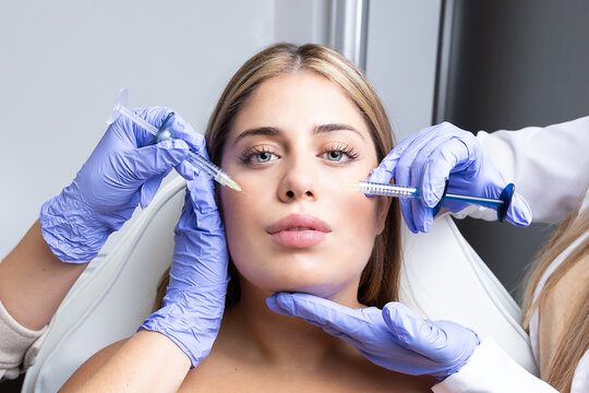 Graceful young female client with long blond hair sitting on medical chair while cut unrecognizable cosmetologists in latex gloves making rejuvenating facial injections procedure in beauty center