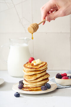 Pouring honey on a stack of fluffy breakfast pancakes with berries and butter
