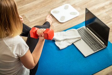 Fototapeta na wymiar Sporty woman using dumbbells making fitness at home via laptop by remote video call online. Young woman losing weight by online gym workout using smart scales.