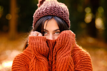 Young female in knitted hat covering face with warm sweater while standing in forest in cold autumn day
